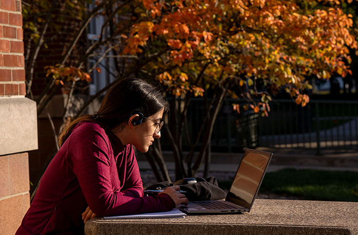 female student sitting at outdoor table studying on laptop. fall foliage in the background.