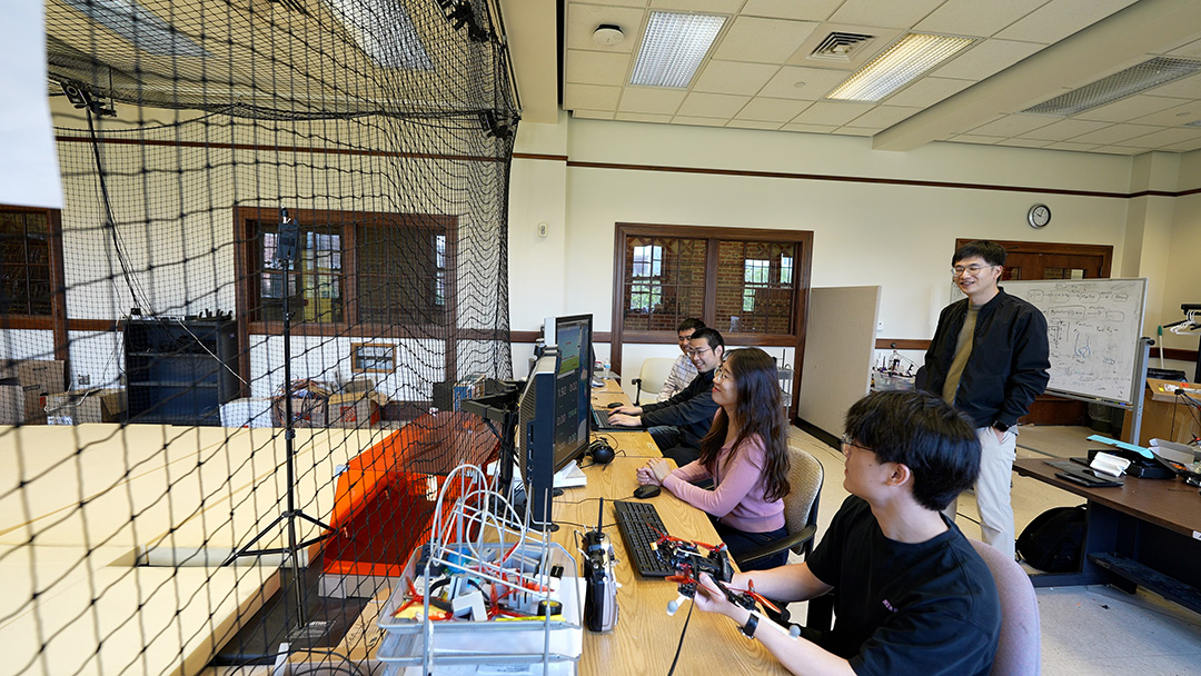 Grad students in the lab, some on the computer, some standing looking at a drone space.