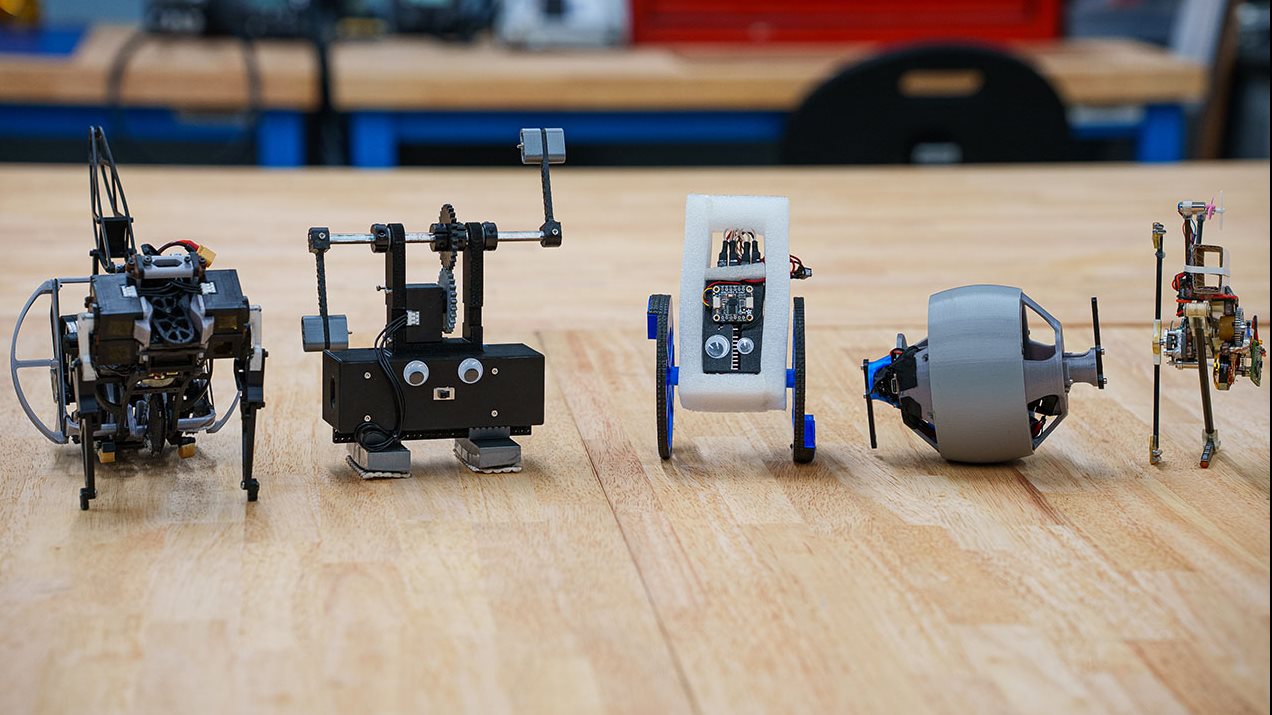 lineup of small robots with googly eyes. Some with legs, some with wheels