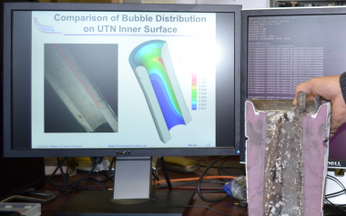 The computer screen shows a computer simulation of the argon gas bubble distribution entering a continuous steel-casting nozzle, which matches the distribution seen in a photo of a water model of the process.  Without proper argon injection, the foreground shows how a nozzle taken from commercial operation can become clogged with solidified steel and inclusions.