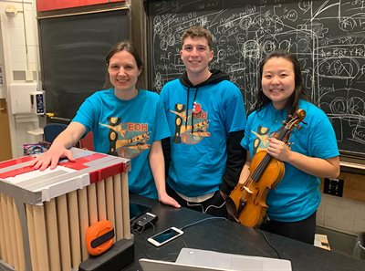 Three students smiling at the camera holding musical instruments in a classroom.