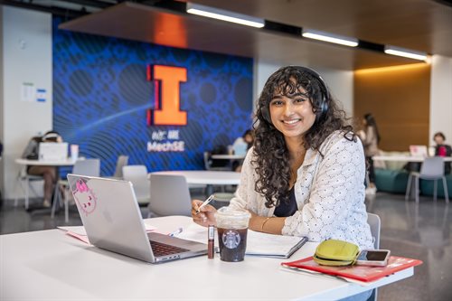 woman student studying at a table with a laptop, smiling at the camera